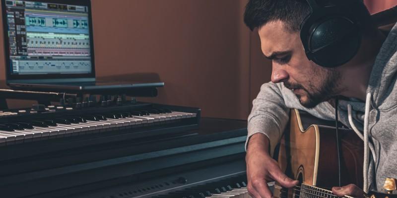 Άνδρας μουσικός συνθέτει μελωδίες παίζοντας κιθάρα σε studio με πλήκτρα και υπολογιστή